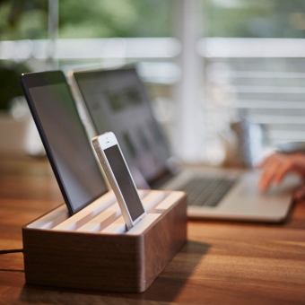 Mobiltelefon und Tablet in Holz-Ladestation und Laptop am Holztisch vor Fenster; bildliche Darstellung des Design-Kompetenzbereichs für mobile Geräte; Farben: silberne Geräte auf hellem Holz vor grünem Naturhintergrund im Fenster